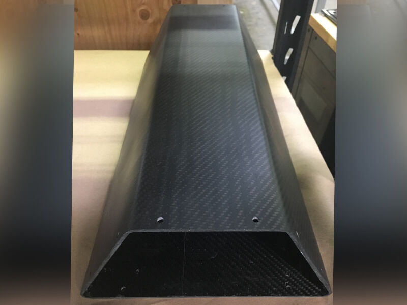 Carbon fiber sensor enclosure laying on a flat surface in an office space. The enclosure has a trapezoidal cross section and machined holes near the end. The surface is a matte carbon fiber weave.